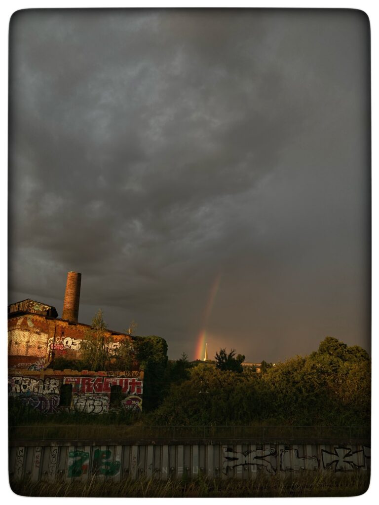 Half a rainbow glowing in a dark, cloudy sky. A sun-lit train yard is in the foreground.