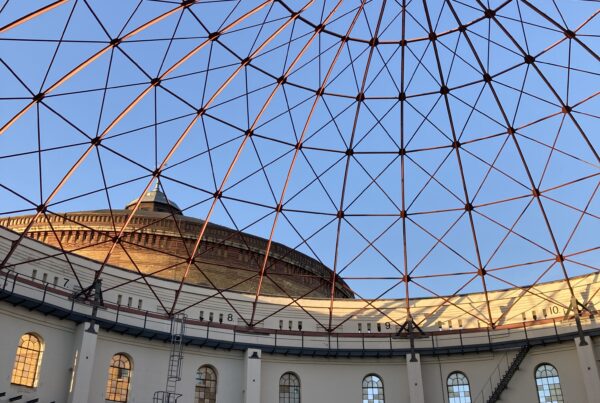 A view from inside a decommissioned gas storage building in Leipzig, looking up through the skeletal iron dome roof to the blue sky above.