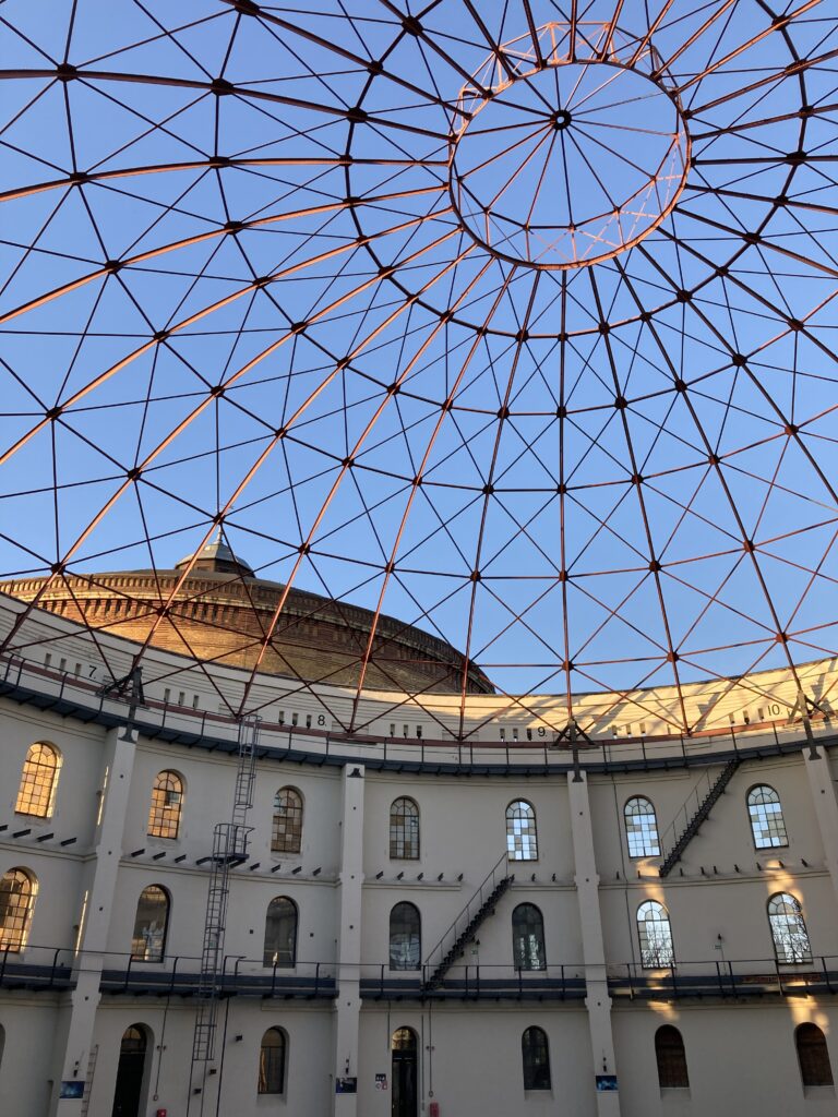A view from inside a decommissioned gas storage building in Leipzig, looking up through the skeletal iron dome roof to the blue sky above.