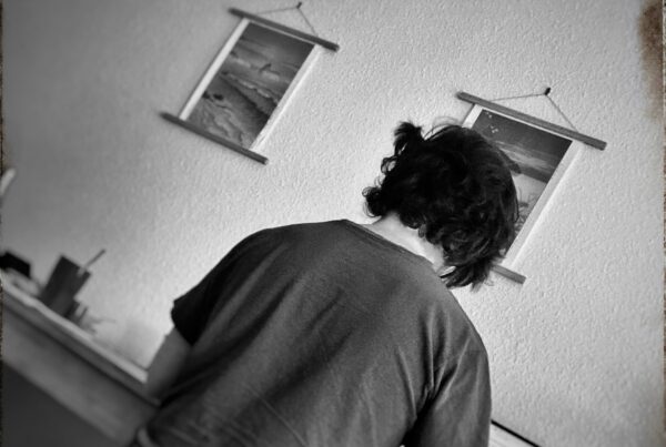Man sitting at a desk indoors working on a laptop. Two pictures hang on the wall.