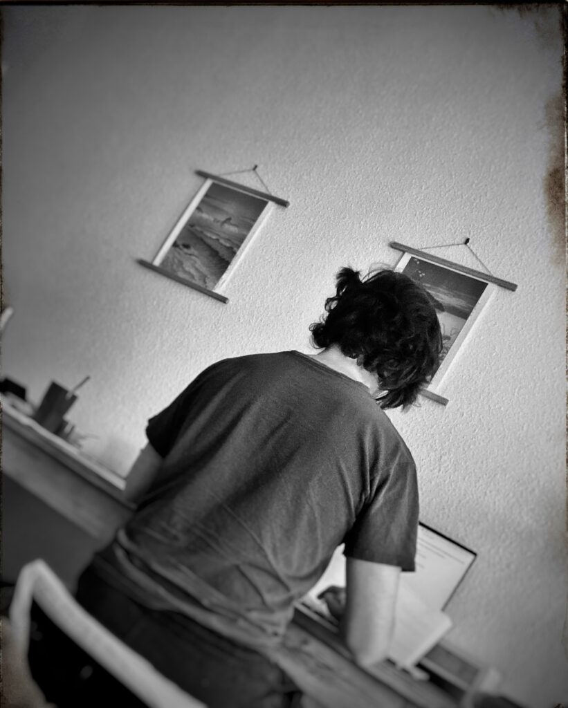 Man sitting at a desk indoors working on a laptop. Two pictures hang on the wall.