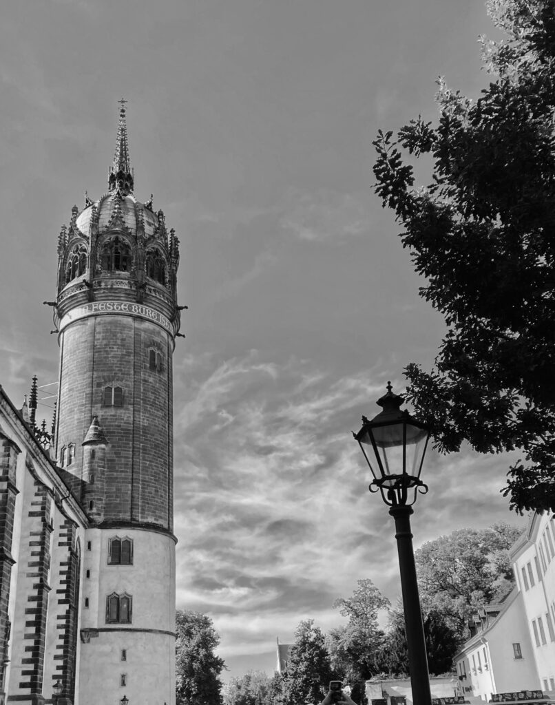A view of the All Saints castle church tower in Lutherstadt-Wittenberg, rising high over the courtyard and the smaller buildings opposite.