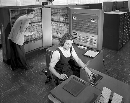 Black and white photo of a man and a woman working by an IBM 700 mainframe computer. She's sitting at an operating terminal, he's standing looking at the mainframe itself.