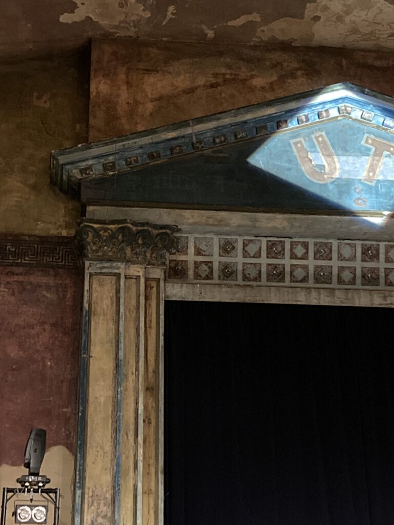 A cropped photograph of a 1920s cinema stage showing one column and a crosspiece bearing the letters UT highlighted by a spotlight (red on blue background).