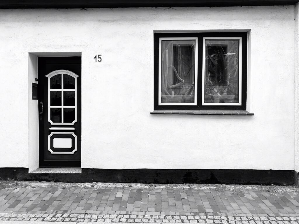 A black and white photo of the front of a house. A door with a 6-segment window is on the left, and a window with cacti behind the panes is on the right. In front of the house are paving tiles borderied by small, neat cobbles.