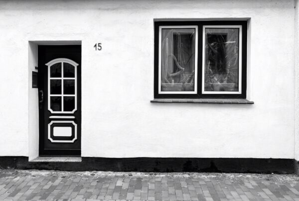 A black and white photo of the front of a house. A door with a 6-segment window is on the left, and a window with cacti behind the panes is on the right. In front of the house are paving tiles borderied by small, neat cobbles.