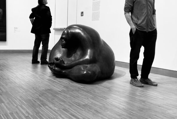 Black and white photo of two men standing besides a large sculpture of an Orangutan sitting on the floor in a meditative pose, from Iris Sageser's Buddhaminders, N-O-W series.