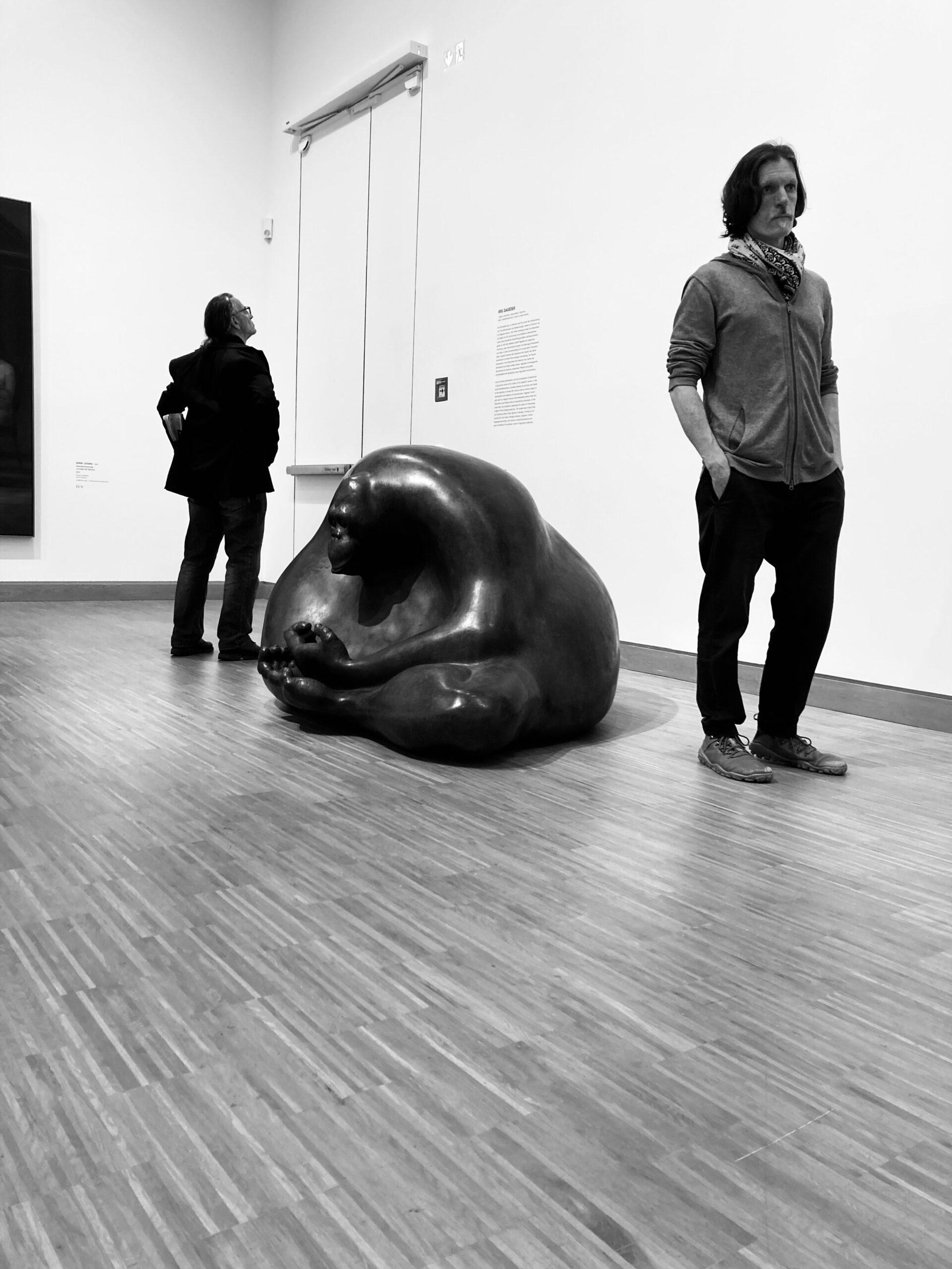 Black and white photo of two men standing besides a large sculpture of an Orangutan sitting on the floor in a meditative pose, from Iris Sageser's Buddhaminders, N-O-W series.
