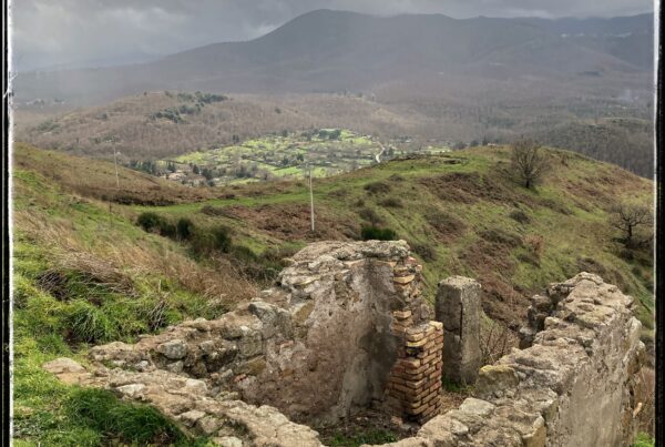 A view from the ruins of Tusculum, on Tuscolo hill near Frascati, Italy. Four crumbling stone walls emerge from the earth in the foreground. In the distance is the green, fertile landscape of the caldera of the dormant Alban volcano. In the distance, a storm brews over the Alban hills.