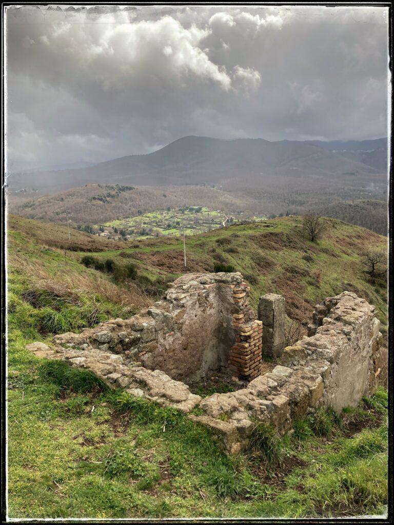 A view from the ruins of Tusculum, on Tuscolo hill near Frascati, Italy. Four crumbling stone walls emerge from the earth in the foreground. In the distance is the green, fertile landscape of the caldera of the dormant Alban volcano. In the distance, a storm brews over the Alban hills.