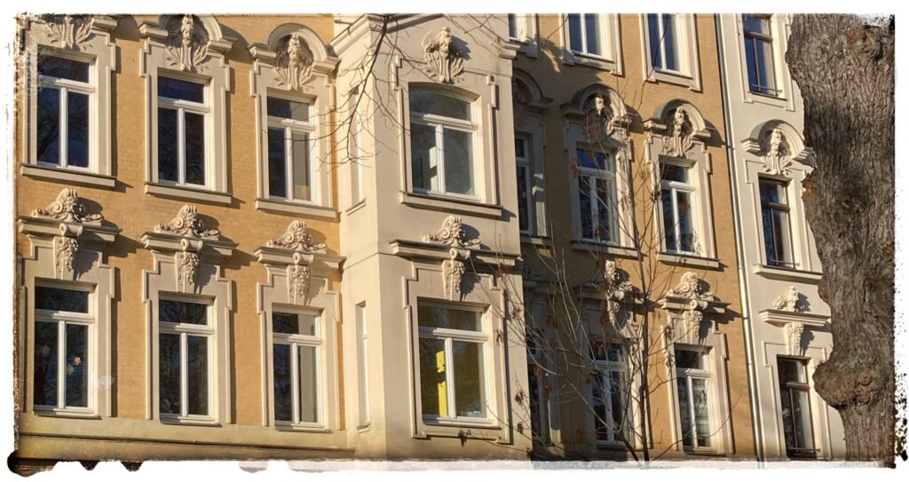 A close up of some windows of a Mehrfamilienhaus showing sculptures of bearded heads above windows, below which hang some grapes.
