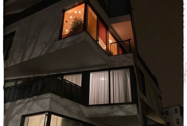 Night image of a block of flats in Leipzig whose every floor is rotated 90 degrees, making the building look like a played-with Rubik's Cube. Soft orange light shines from a flat on the third floor, and there's a decorated Christmas tree in the window.