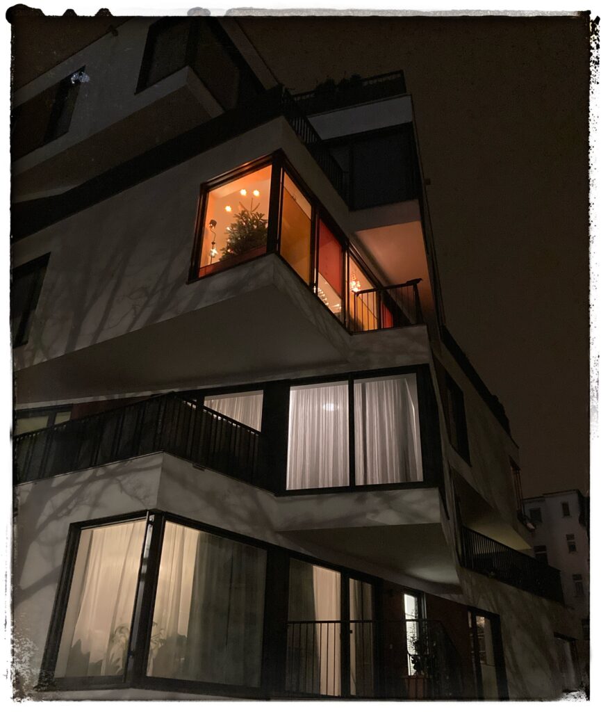Night image of a block of flats in Leipzig whose every floor is rotated 90 degrees, making the building look like a played-with Rubik's Cube. Soft orange light shines from a flat on the third floor, and there's a decorated Christmas tree in the window.