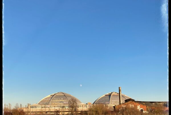 A photo of two giant, ribbed concrete domes reflecting the setting sun. A full moon rises between them. In the foreground is an area of brown-green heathland. In the background, a cloudless blue sky.