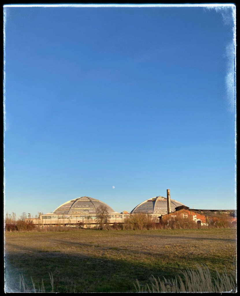 A photo of two giant, ribbed concrete domes reflecting the setting sun. A full moon rises between them. In the foreground is an area of brown-green heathland. In the background, a cloudless blue sky.