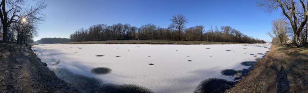 Panoramic photo of the frozen river Elster, Leipzig. The ice is covered in snow, save for a few mysterious clear circles. On the far bank, a line of trees is visible. The sky is clear blue.