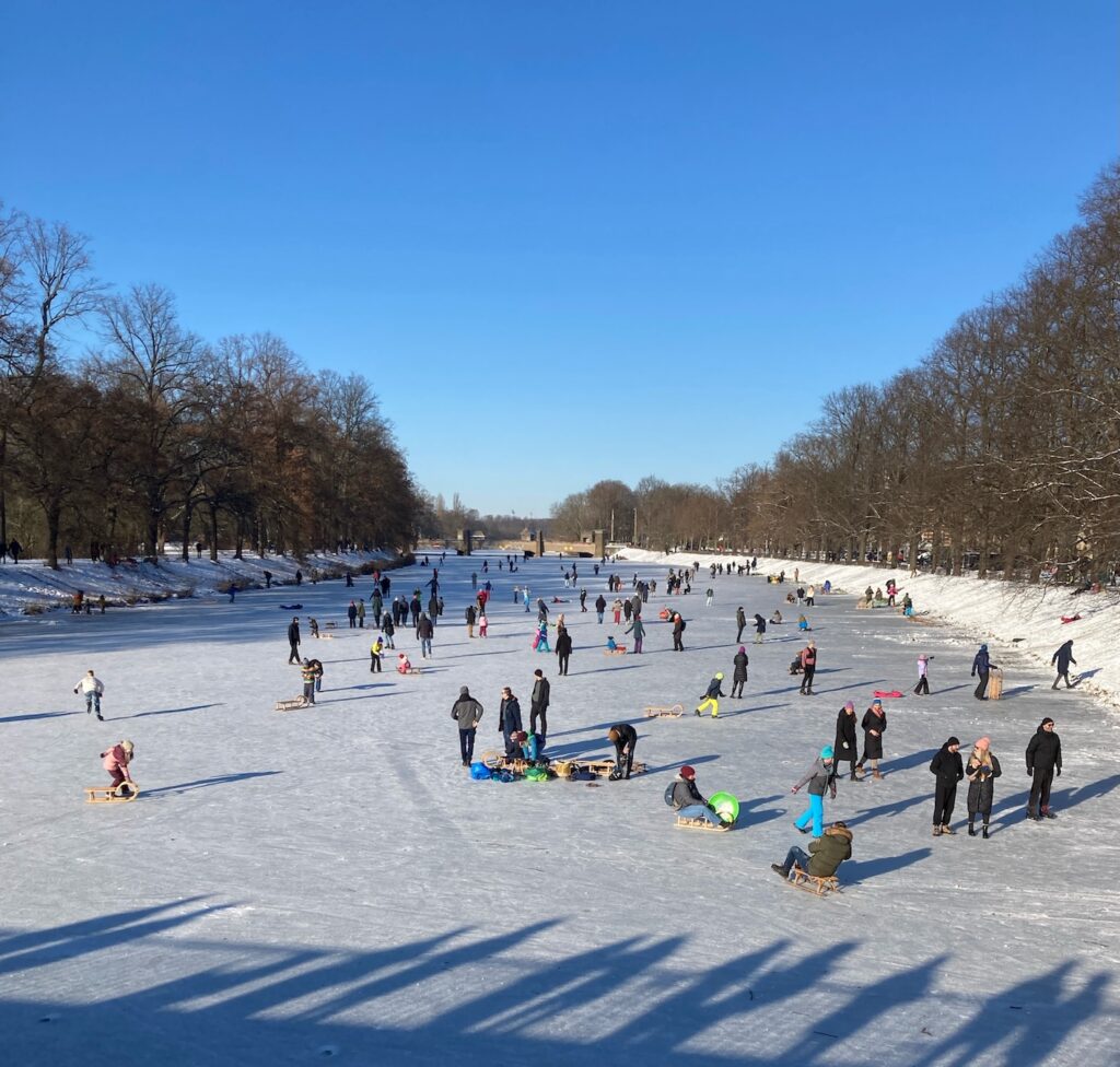 A photo of the river Elster in Leipzig, frozen solid. The river stretches away into the distance, with tree-lined banks on either side. Families are out on the ice. Children are playing on sledges, grown-ups are walking and sliding along.