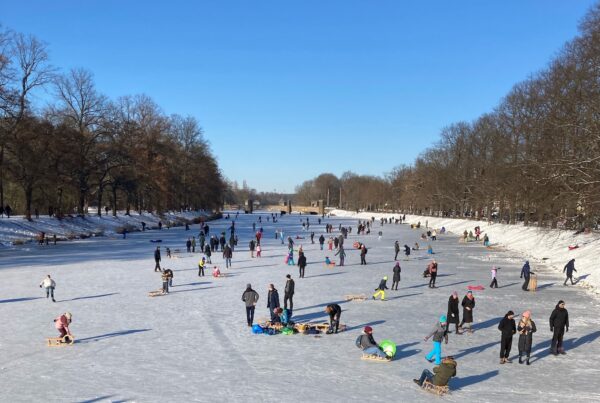 A photo of the river Elster in Leipzig, frozen solid. The river stretches away into the distance, with tree-lined banks on either side. Families are out on the ice. Children are playing on sledges, grown-ups are walking and sliding along.