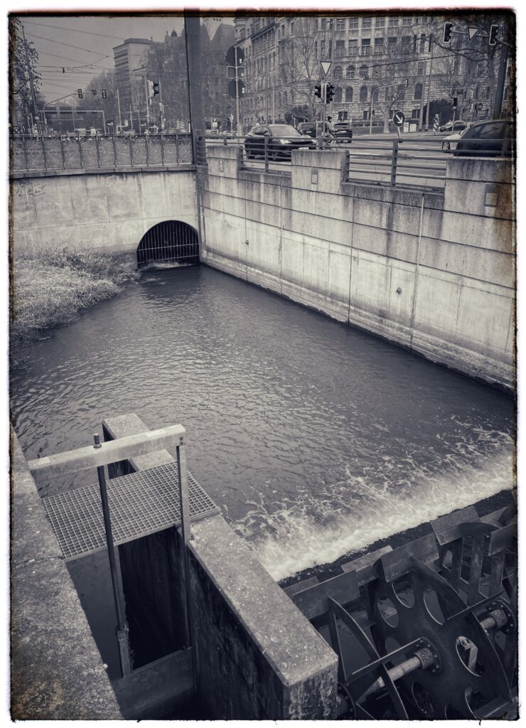 Photo of a stretch of the old Pleiße mill stream in Leipzig. In the foreground is a stationary hydraulic roller, marking where an old paper mill used to be. In the background, the stream disappears into a tunnel than runs beneath a busy crossroad.
