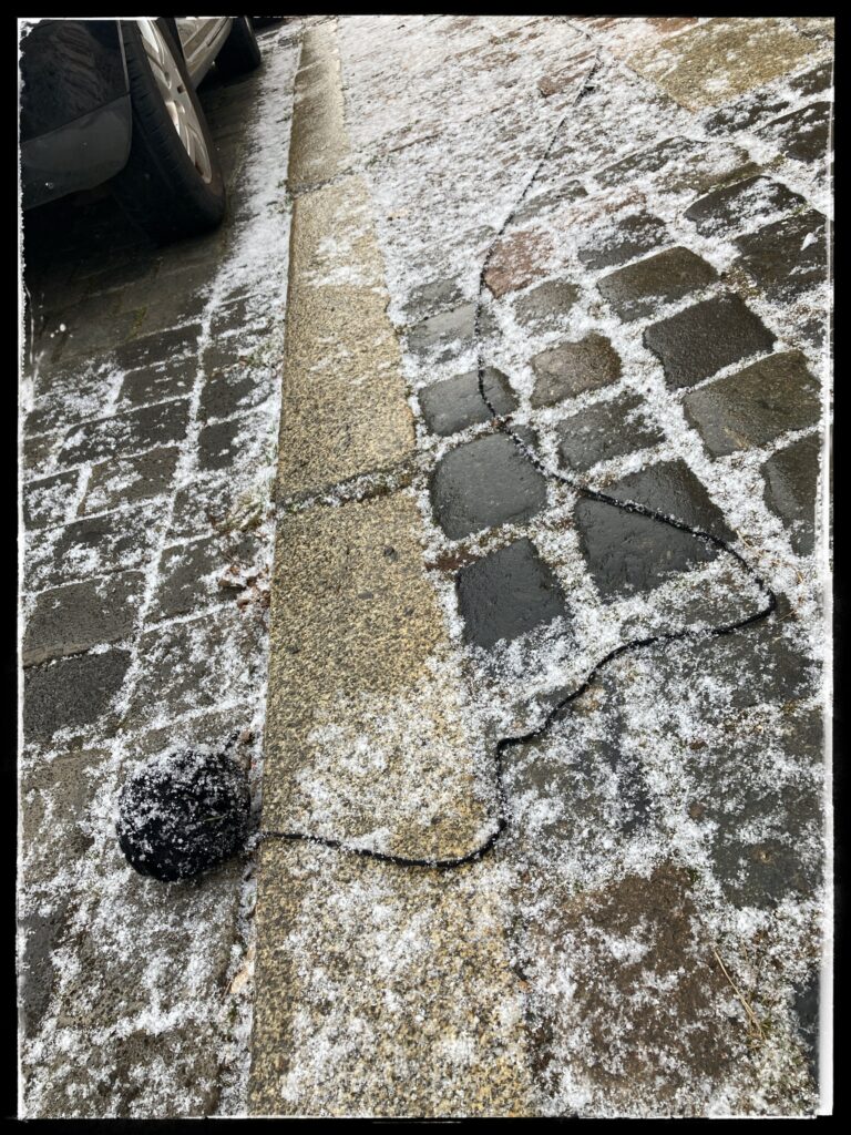 Photo of a ball of black wool lying on a road by a kerb. It's partly unravelled. A line of black yarn trails off along the pavement.
